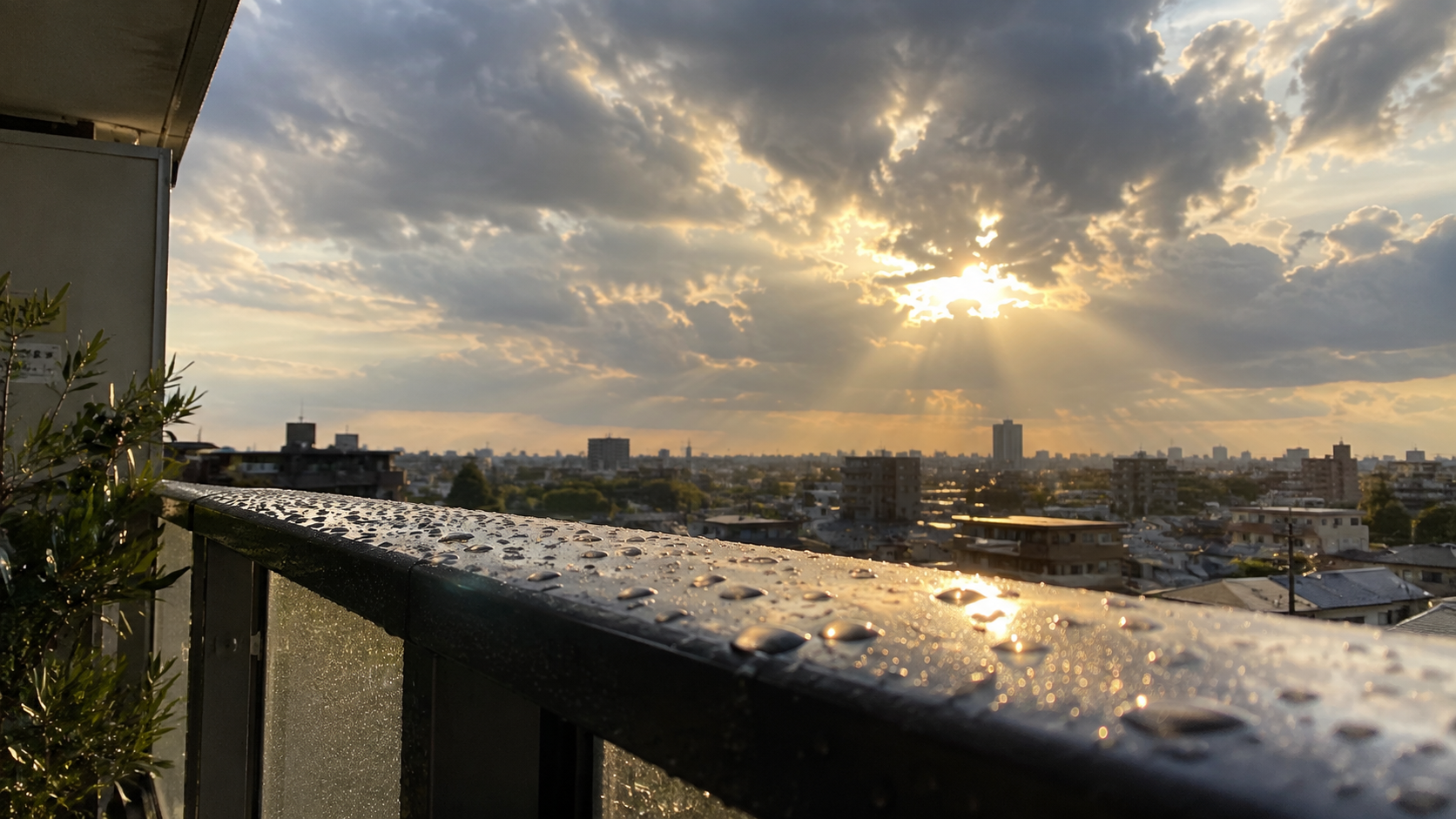 水滴が残るベランダの手すりと、雲間から日が差す空